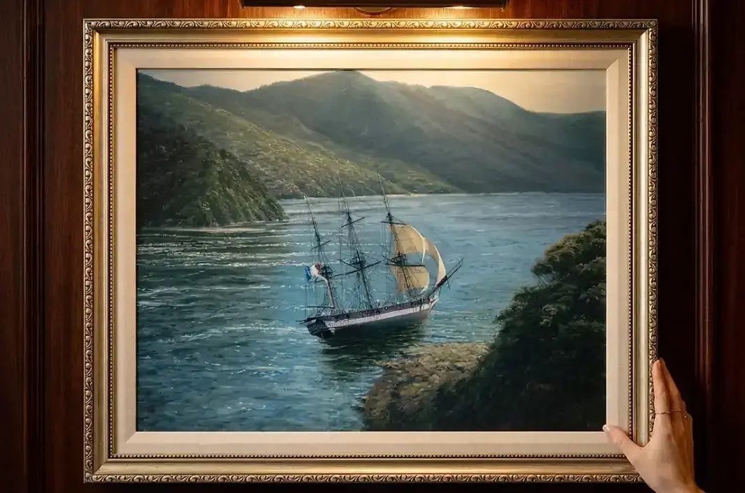 The framed painting held up by two hands against rich dark wooden wall panelling under a warm picture light.