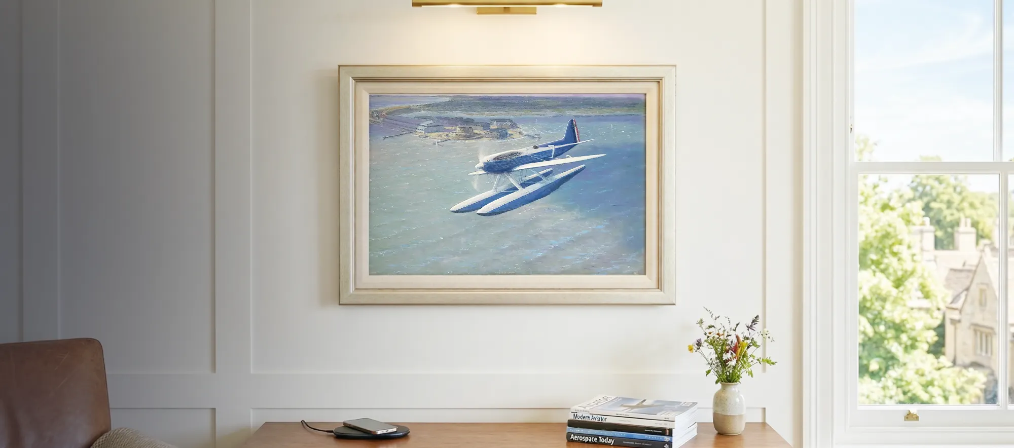 Framed aviation painting hanging in a bright, modern study above a walnut credenza, illuminated by a sleek brass picture light.
