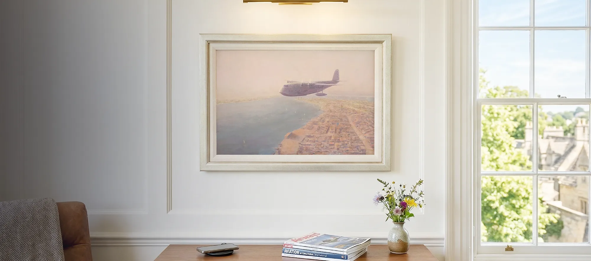 Framed aviation painting hanging in a bright, modern study above a walnut credenza, illuminated by a sleek brass picture light.