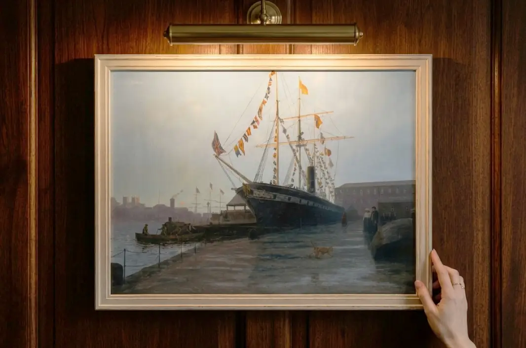 The framed painting held up by two hands against rich dark wooden wall panelling under a warm picture light.