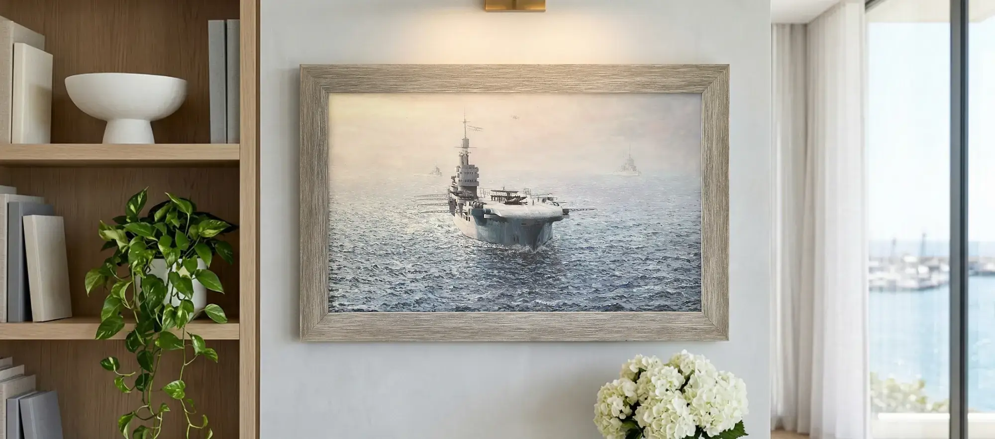 Framed panoramic painting of HMS Illustrious hanging on a white wall above a light wood table with white hydrangeas, flanked by a modern bookcase.