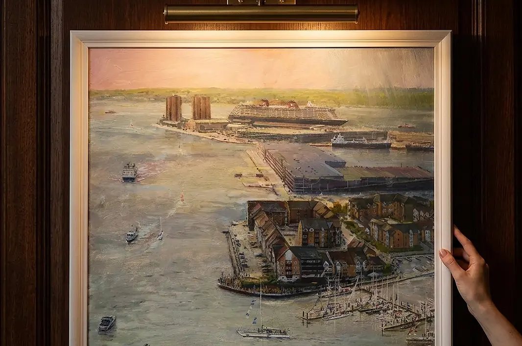 A person's hands adjusting the vertically framed painting against rich dark wooden wall panelling under a warm picture light.