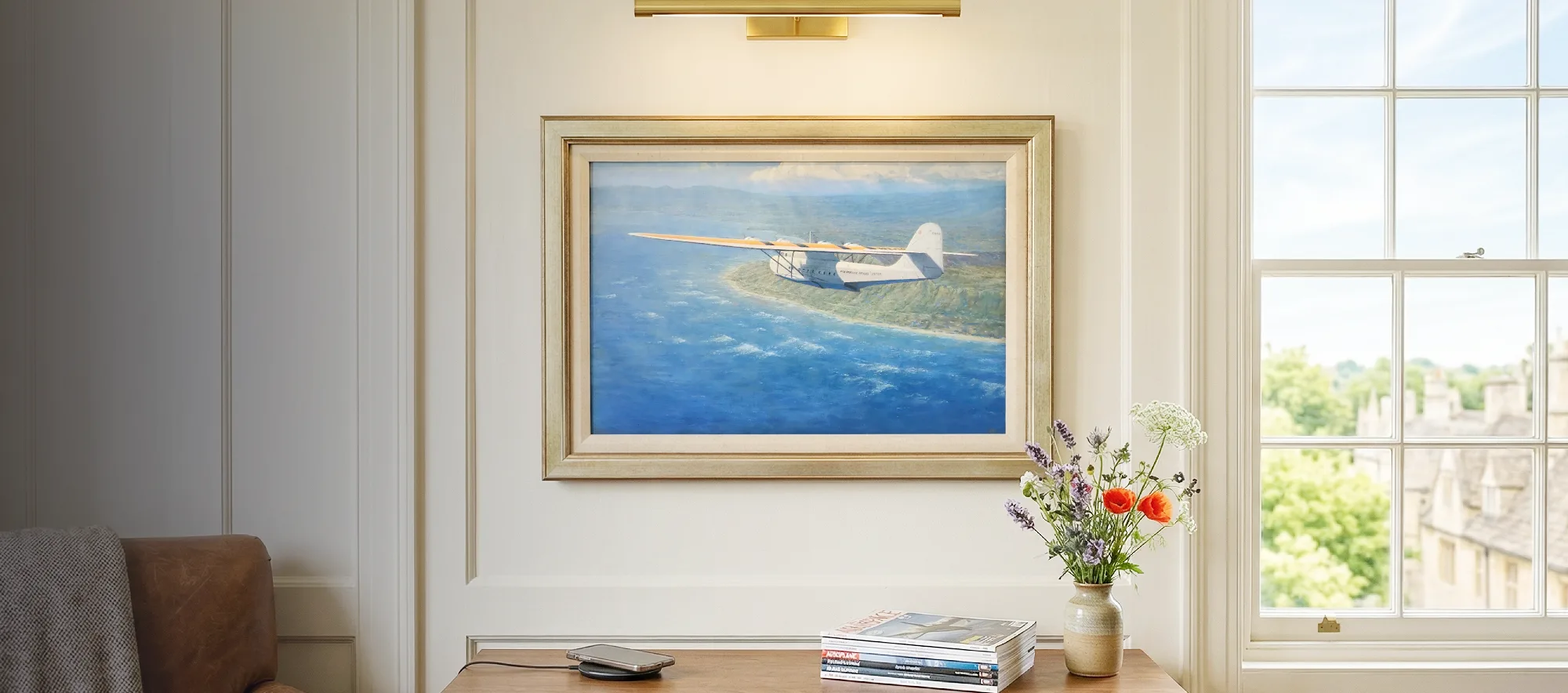 Framed aviation painting hanging in a bright, modern study above a walnut credenza, illuminated by a sleek brass picture light.
