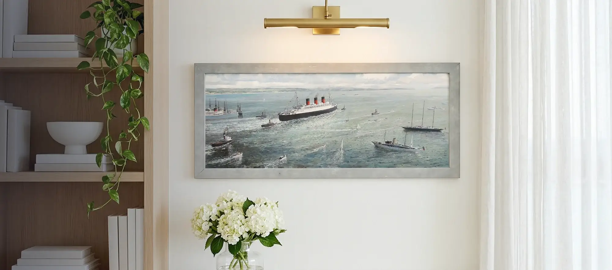 Framed panoramic painting of the Queen Mary hanging on a white wall above a light wood table with white hydrangeas.
