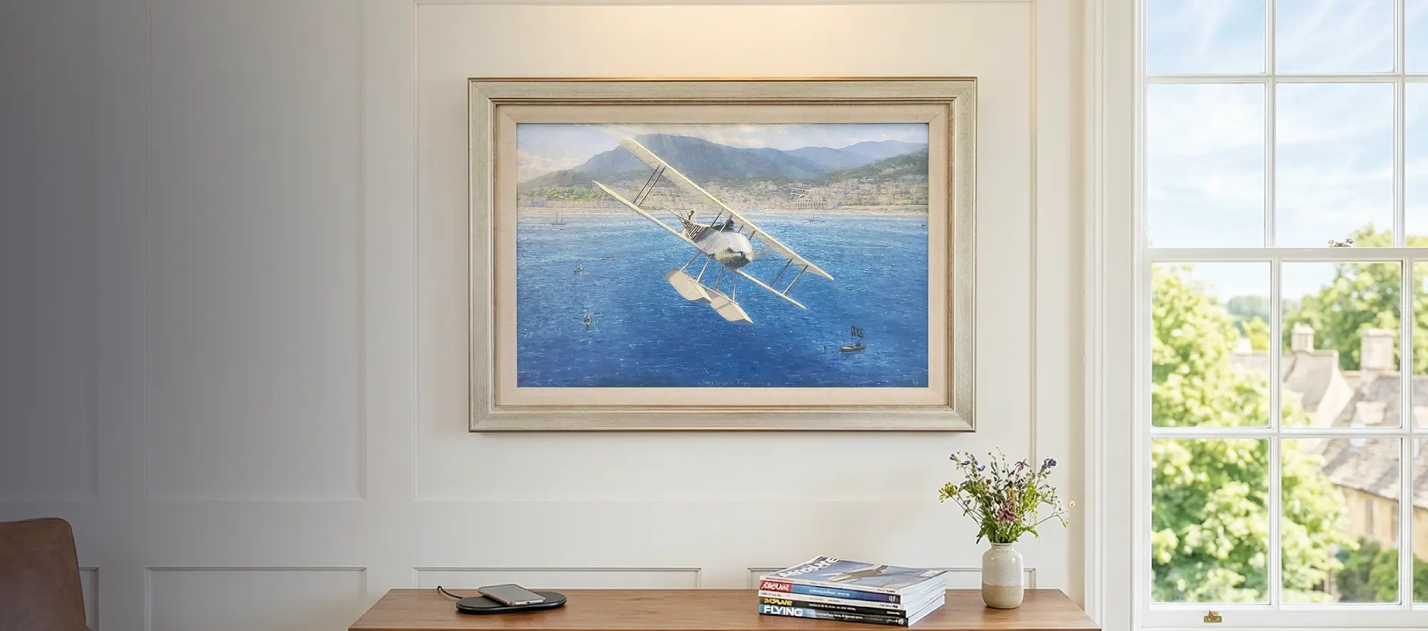 Framed aviation painting hanging in a bright, modern study above a walnut credenza, illuminated by a sleek brass picture light.