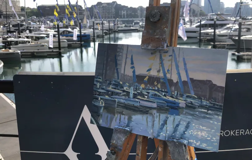 The painting shown on a wooden easel on the dockside, with the actual marina and event signage visible behind it.