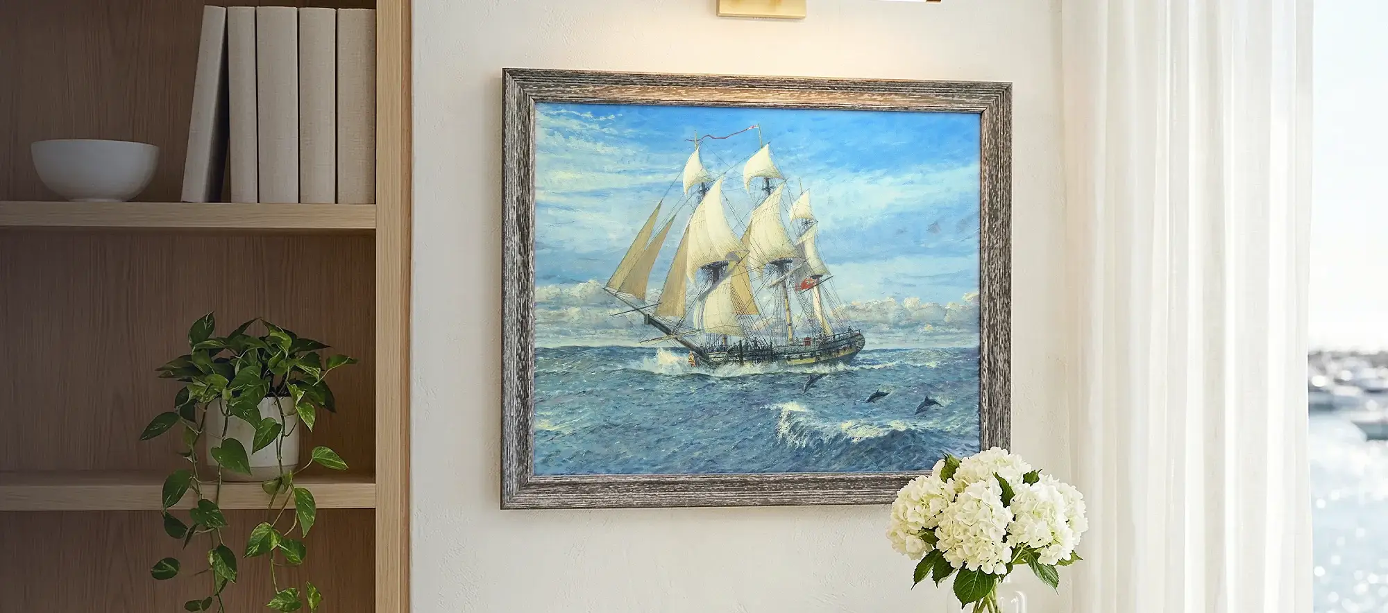 Framed painting of HMS Sphinx hanging on a bright wall above a light wood table with white hydrangeas, flanked by a modern bookcase.