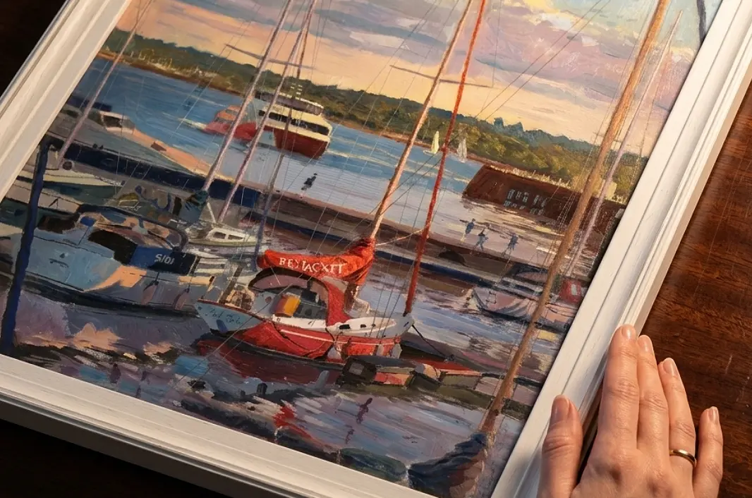 A person's hands adjusting the vertically framed painting against rich dark wooden wall panelling under a warm picture light.