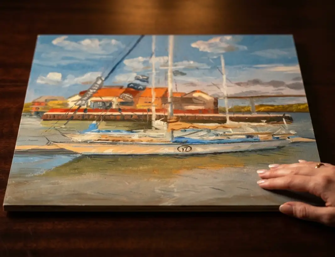 The unframed painted panel held flat against a dark wooden table by a hand.