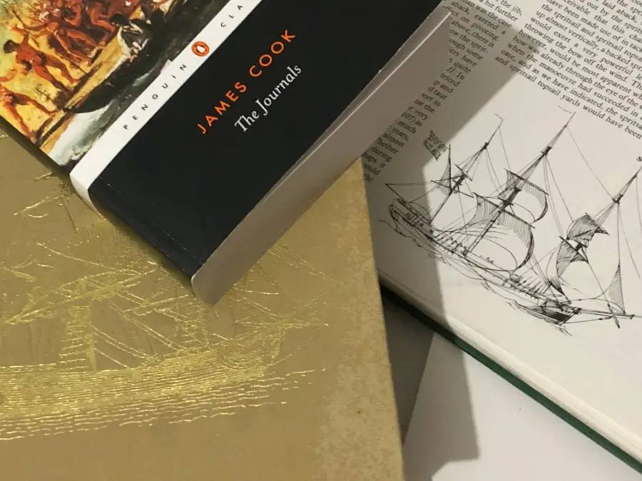 A collection of historical reference materials for a painting commission, featuring the book 'James Cook: The Journals' alongside an open book showing detailed line drawings of a ship's rigging.