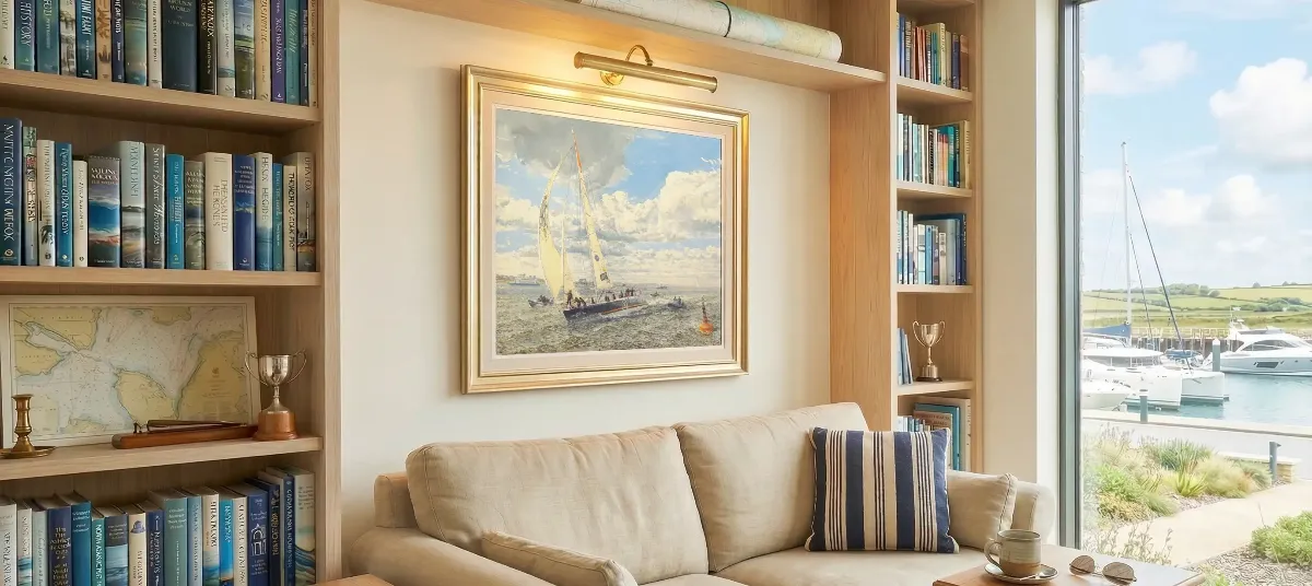 A framed oil painting mounted above a beige sofa and flanked by wooden bookshelves in a nautical-themed room. The painting shows a sailing boat racing under a dynamic, cloud-filled sky, with smaller boats visible in the distance