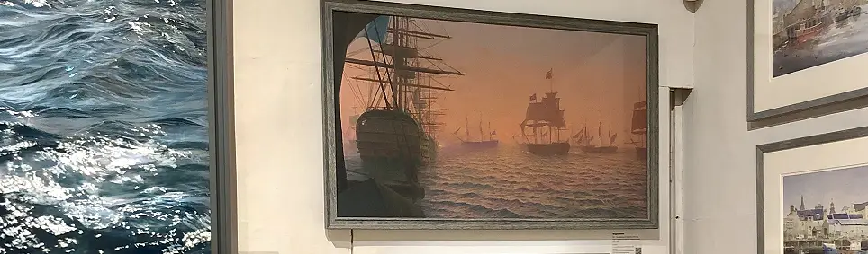 A framed oil painting of a fleet of historical sailing ships silhouetted against a warm, hazy golden sky, hanging on display on a gallery wall at the Royal Society of Marine Artists' annual exhibition 2025.