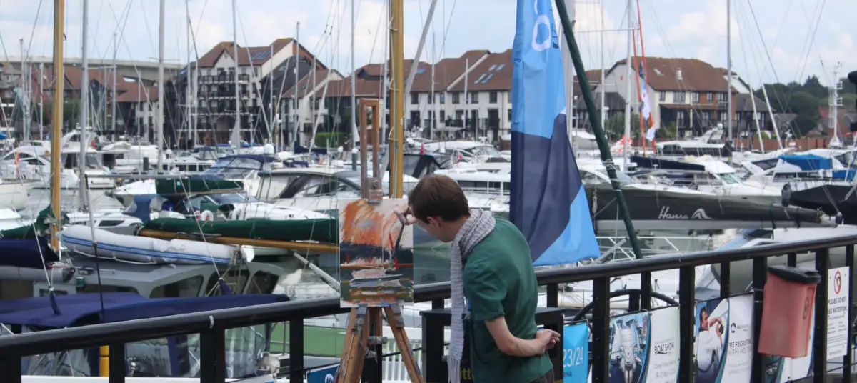 Gregory Smith painting in oils in Ocean Village marina with large numbers of yachts and buildings in the background on a sunny day.